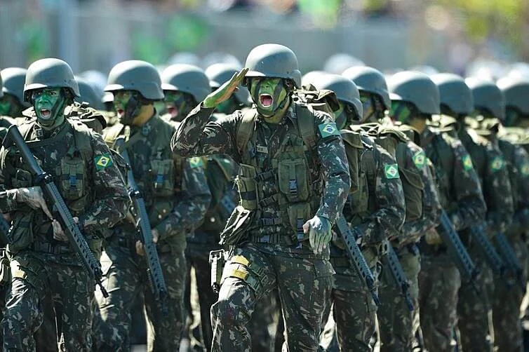 Fotografia de um pelotão de soldados do Exército Brasileiro marchando. Todos usam capacetes verdes, uniformes camuflados com a bandeira do Brasil na manga e pintura de camuflagem verde e preta no rosto. O soldado em destaque no centro está marchando, prestando continência com a mão direita no capacete e com a boca aberta em tom de marcha.